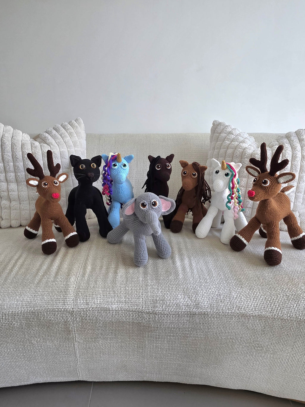 A group of handmade crochet amigurumi animals, including two reindeer, a black cat, a blue and white unicorn, a chocolate and hazel horse and a grey elephant featuring at the front.