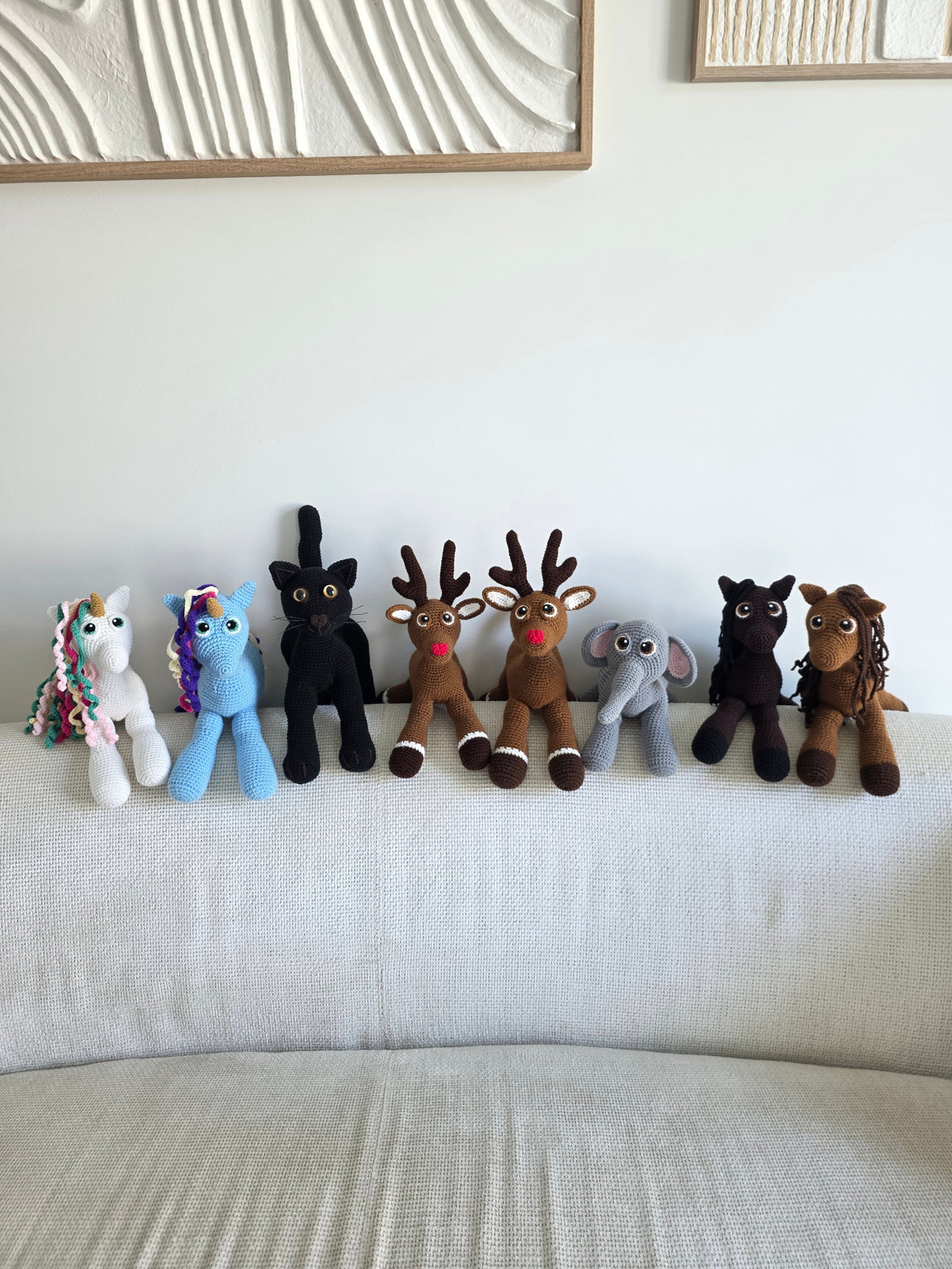 A group of handmade crochet amigurumi animals, including two reindeer, a black cat, a grey elephant, a blue and white unicorn, a chocolate and hazel horse.